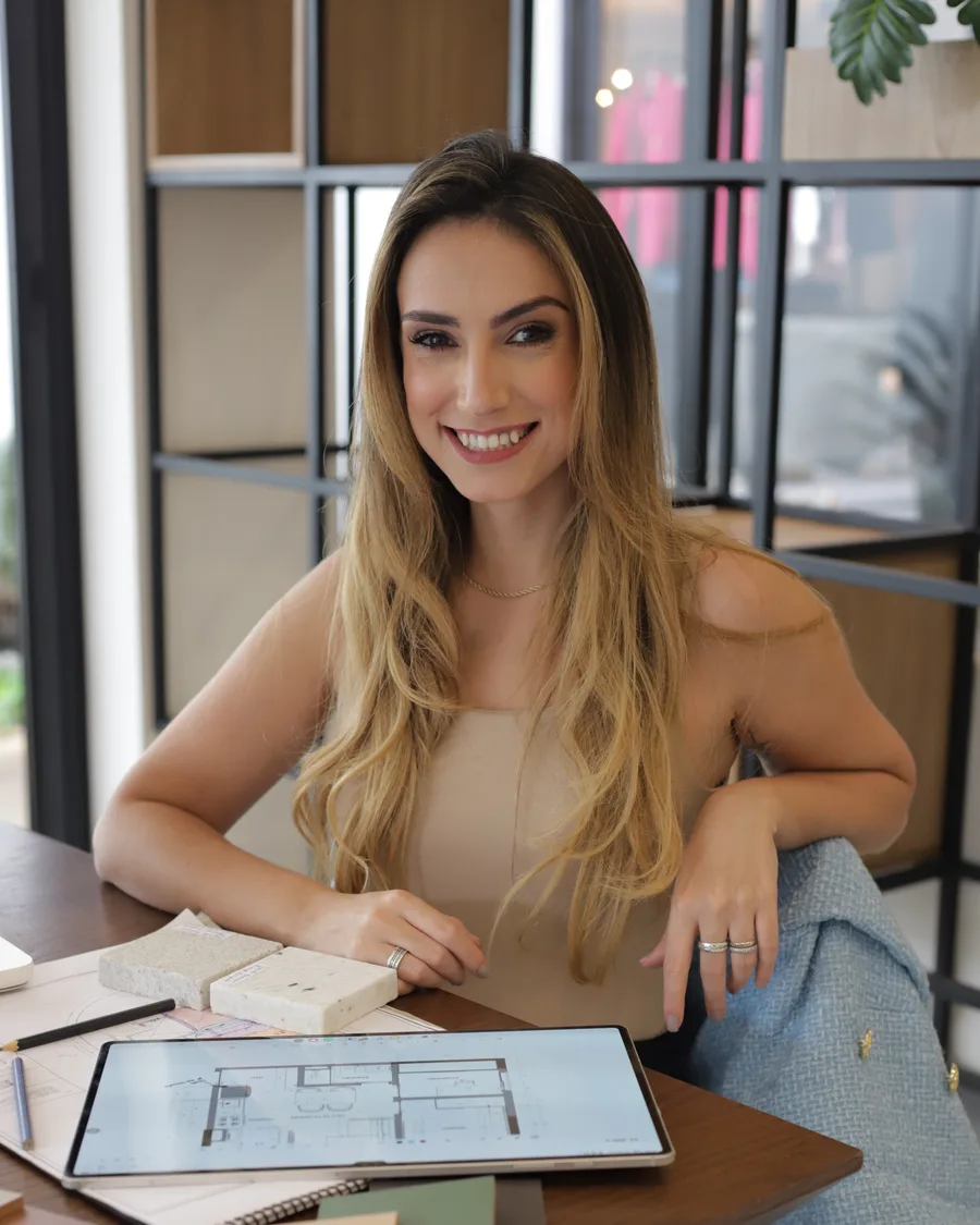 Fernanda Bortoli sorrindo à mesa de trabalho, com planta no tablet e amostras de acabamento